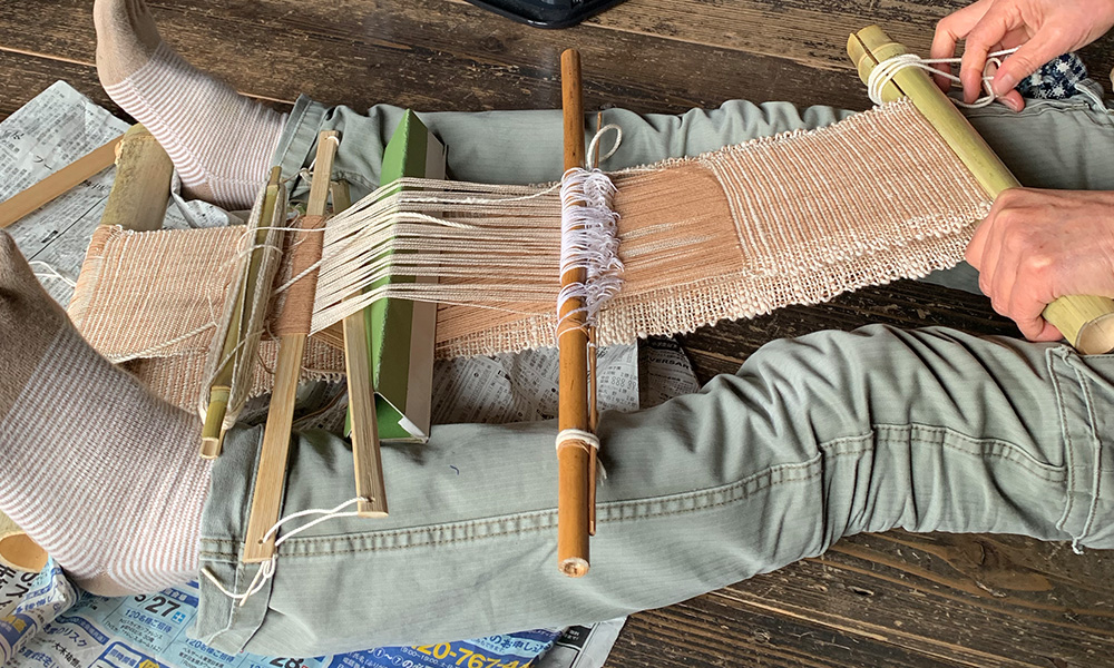 原始ばたを使って織っている様子。今回使う縦糸は藍染した木綿糸を使うので、こちらで織っているものは別のもの。竹の道具等、1点1点手作りしたものを使うので、写真と実物は異なります。